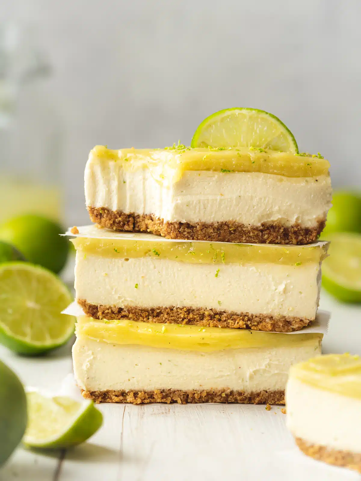 a stack of vegan lime cheesecake bars with a lime wedge on top and fresh limes scattered around. There is a bite taken out of the top slice showing the creamy texture.