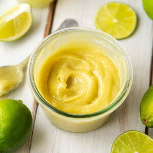fresh homemade lime curd in a glass jar with fresh limes and lime slices scattered around it.