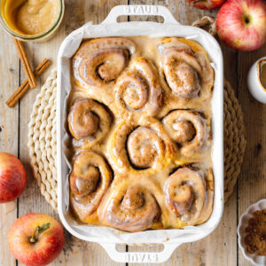 cinnamon rolls with apple pie filling in a staub pan with vanilla glaze. There are fresh gala apples scattered around the scene.