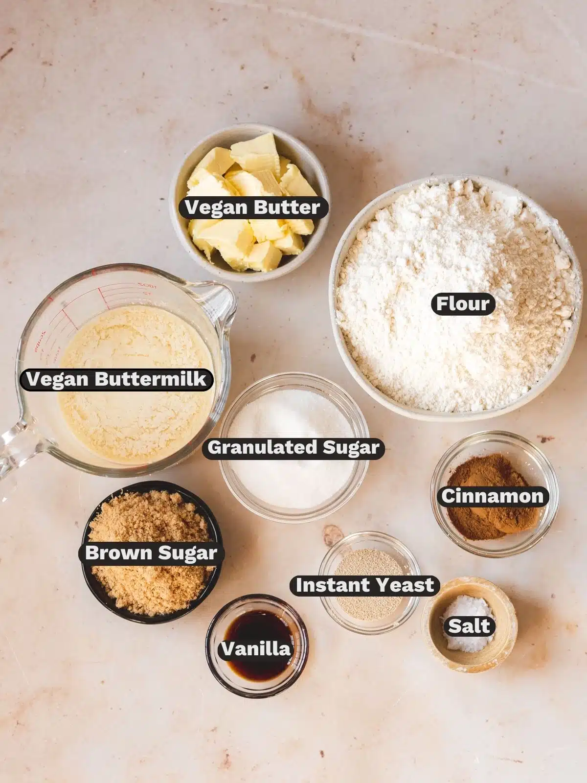 Ingredients needed to make vegan cinnamon buns measured out into bowls on a white table with text overlay.
