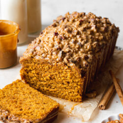 a loaf of vegan pumpkin bread with a slice taken from it showing the fluffy texture, It has a biscoff glaze and pecan streusel topping.