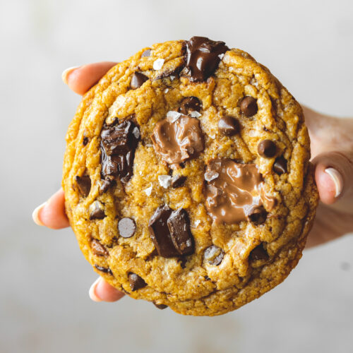 Closeup side on shot of a hand holding a giant vegan pumpkin chocolate chip cookie showing the puddles of melted chocolate on top.