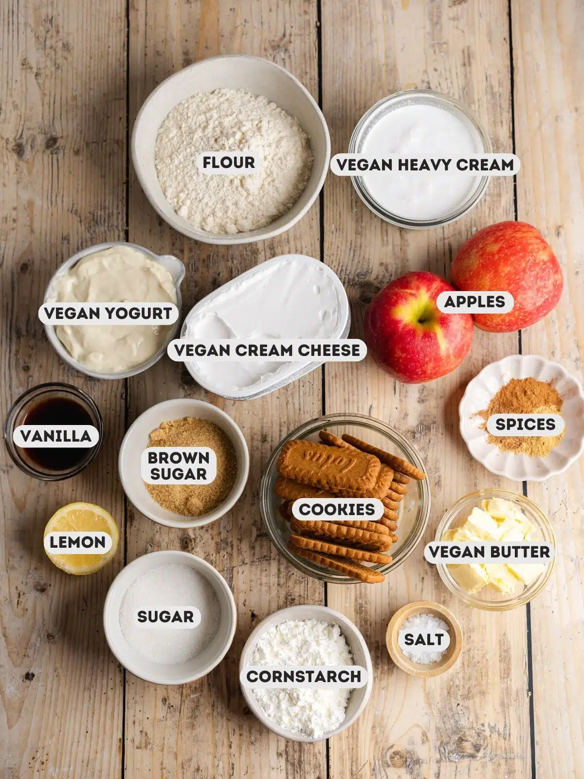 Ingredients needed to make a vegan apple crumb cheesecake measured out into bowls on a wooden table with text overlay.
