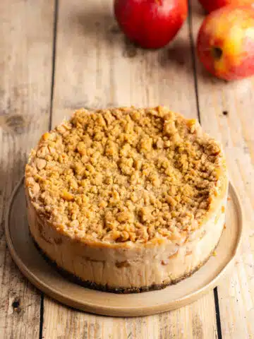 Baked apple crumble cheesecake on an earthenware plate on a wooden table with some whole apples.