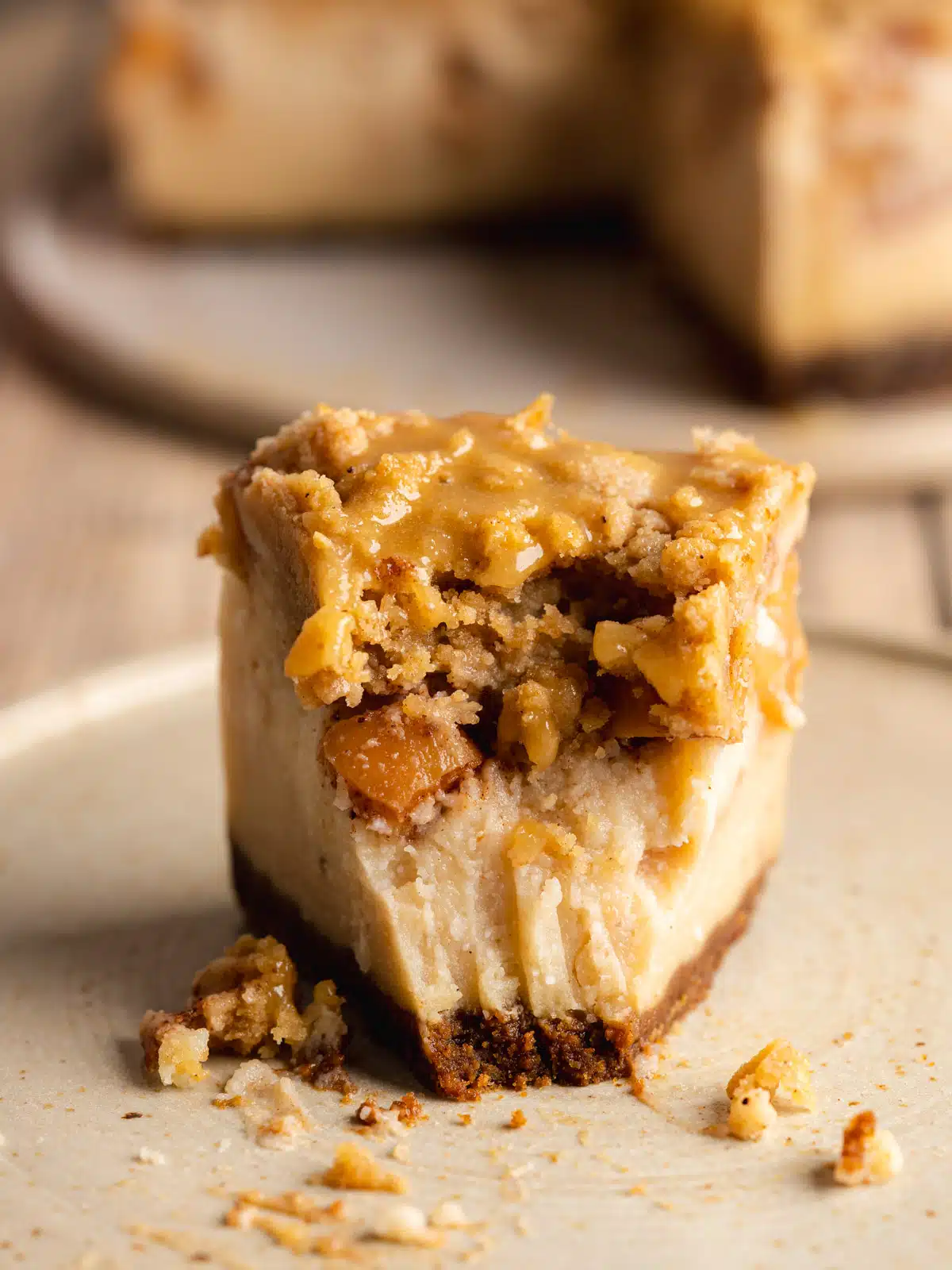 Slice of apple crumble cheesecake on a plate with the pointy end facing the camera and a bite taken out to show the 4 distinct layers of biscoff crust, creamy cheesecake filling, spiced apples, and crumb topping.