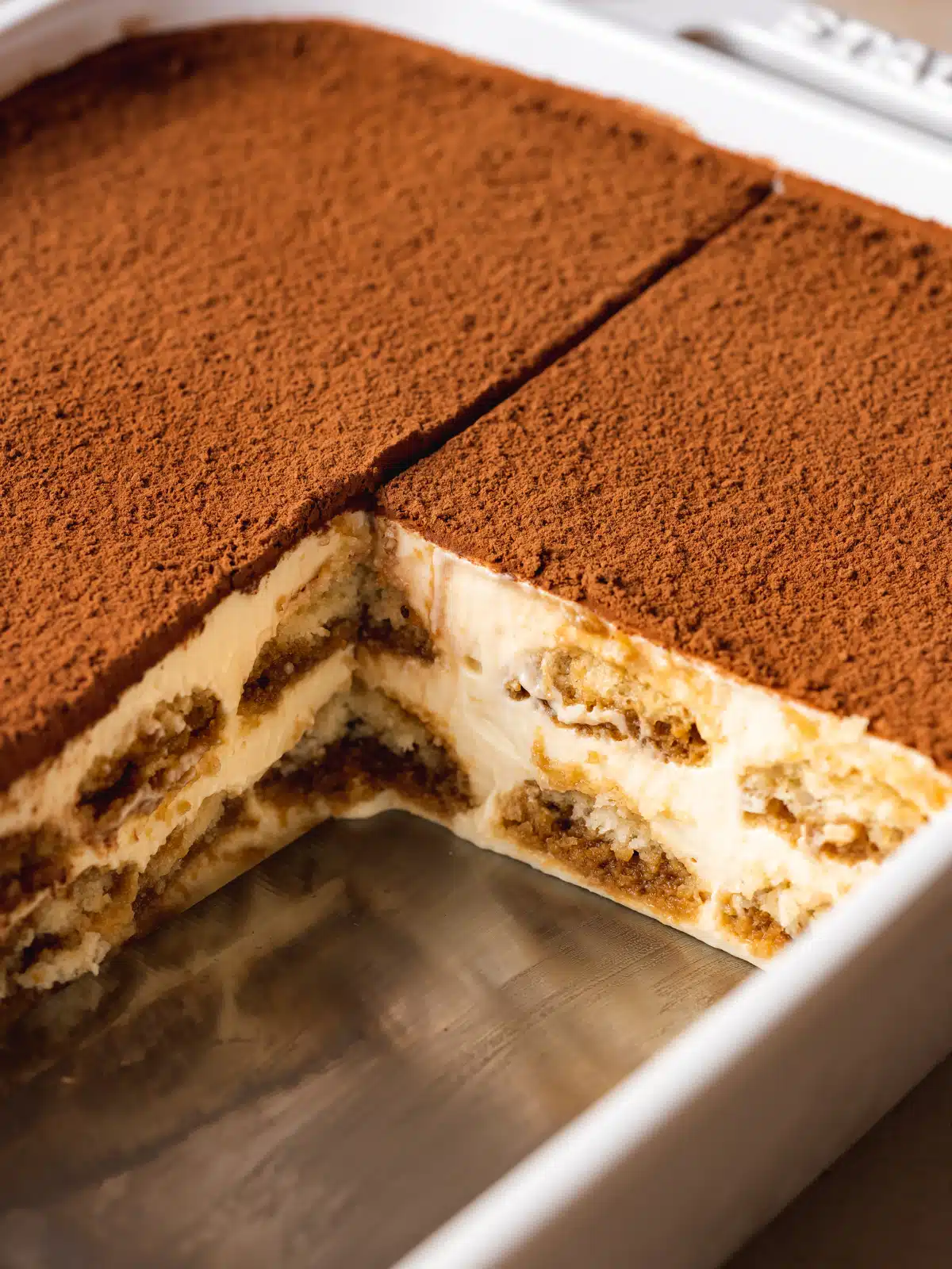 perfectly clean cuts of tiramisu with a slice missing. Shows a cross-section of espresso rum soaked ladyfingers between layers of mascarpone cream.