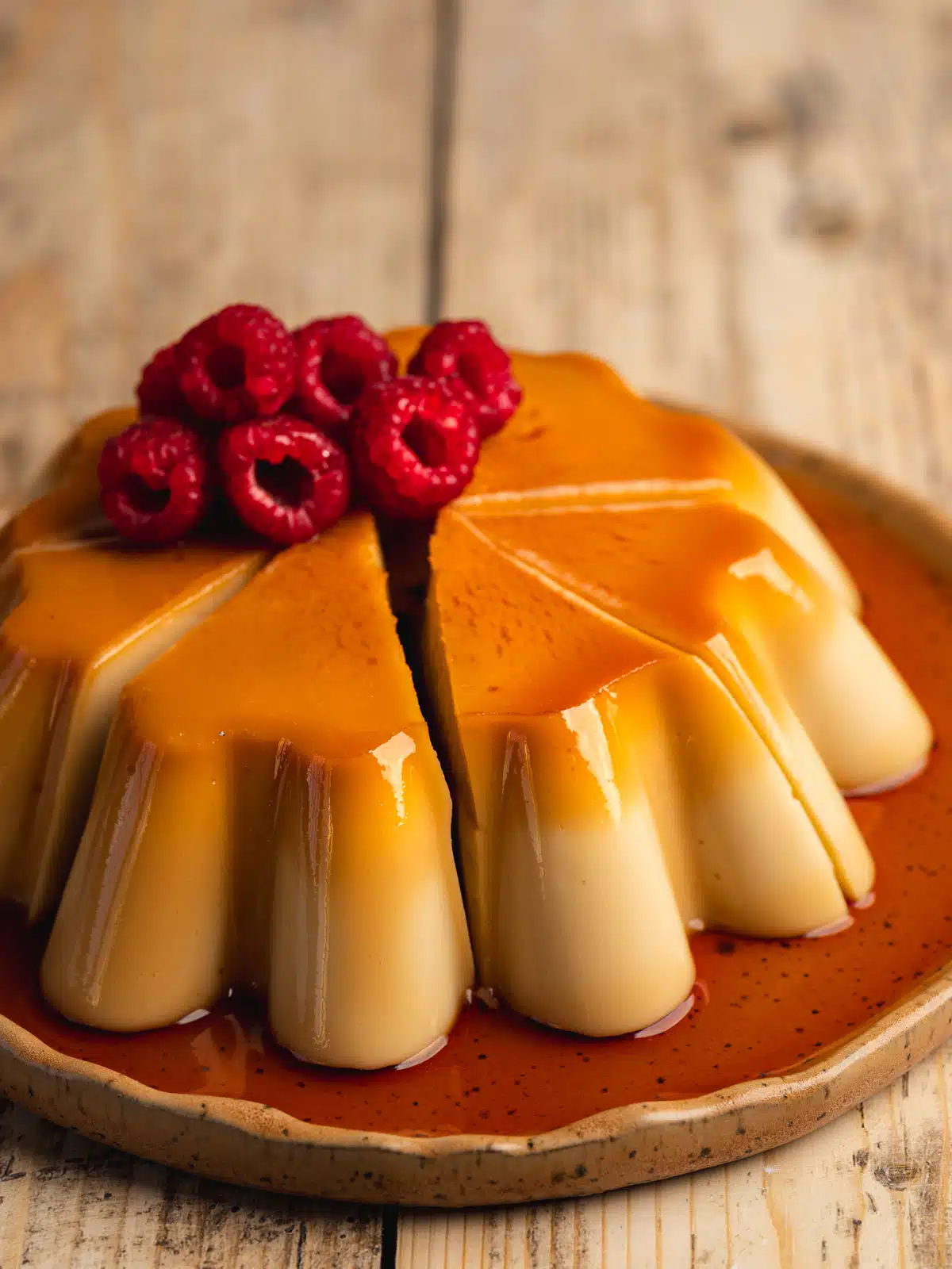 a vegan caramel flan topped with raspberries and caramel oozing over the edges on an earthenware plate.