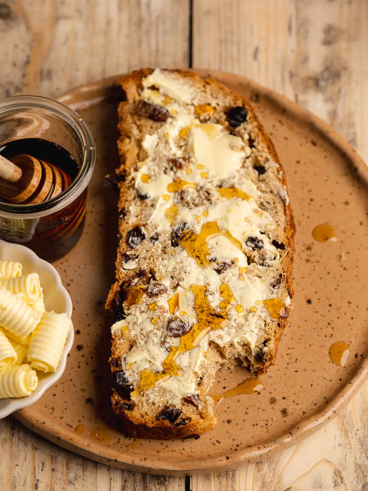 a slice of buttered barmbrack on an earthenware plate with a drizzle of maple syrup on top and a bite taken out of it.