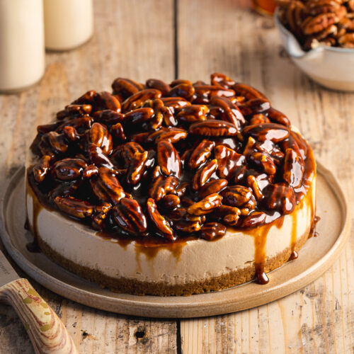 a no bake vegan cheesecake topped with pecan pie topping with caramel oozing down the sides on top of a ceramic plate.