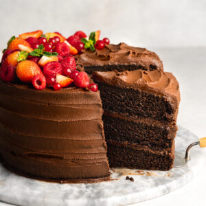 Side on view of a 3-layer chocolate frosted vegan chocolate cake topped with berries on a marble cake stand with a cake server removing a slice.
