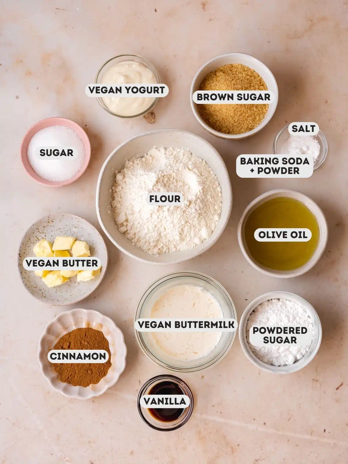 Overhead shot of the ingredients needed to make vegan cinnamon streusel muffins measured out into bowls on a tan table with text overlay.