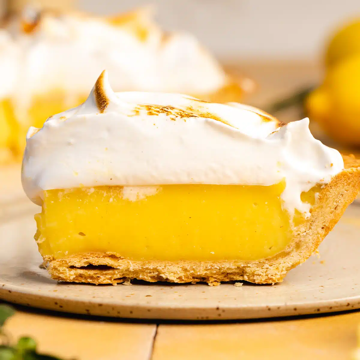 a close up side shot of a slice of lemon meringue pie showing a flaky crust and tall layers of bright yellow lemon curd filling and torched meringue topping.