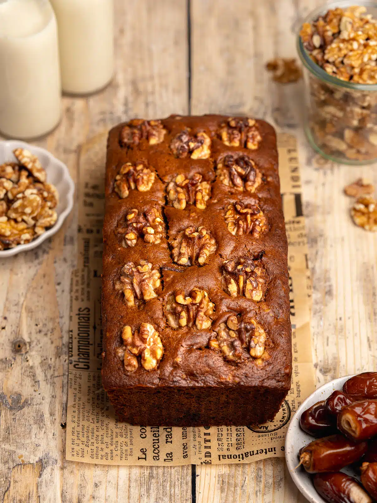 a loaf of freshly baked date cake with walnuts on top on a wooden surface with whole dates and walnuts scattered around.