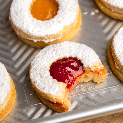 apricot and strawberry jam filled linzer cookies on a metal baking sheet, with a bite taken from one of the cookies.