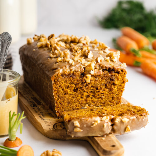 a loaf of carrot cake with brown butter glaze and chopped walnut topping on a wooden cutting board with fresh carrots in the background.