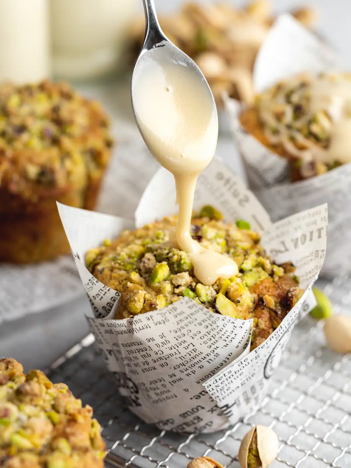 pistachio muffins with streusel topping in tulip muffin liners with a spoon drizzling vanilla sugar glaze on top.