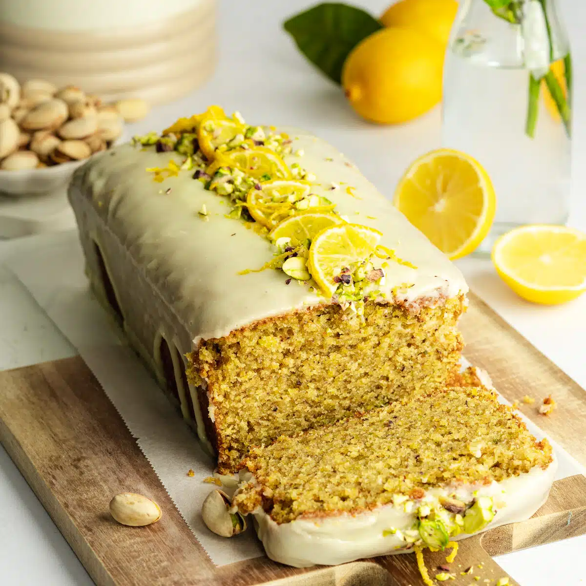 a lemon olive oil pistachio cake decorated with cream cheese, lemon slices and pistachios, there is a slice taken from it revealing the tender interior of the cake.