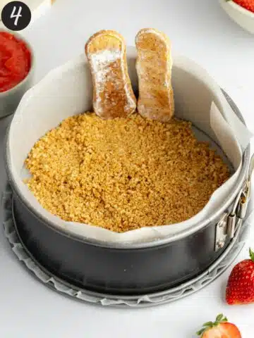 a cake pan lined with parchment paper with cookie crumbs in the base and two ladyfinger cookies lined around the edge.