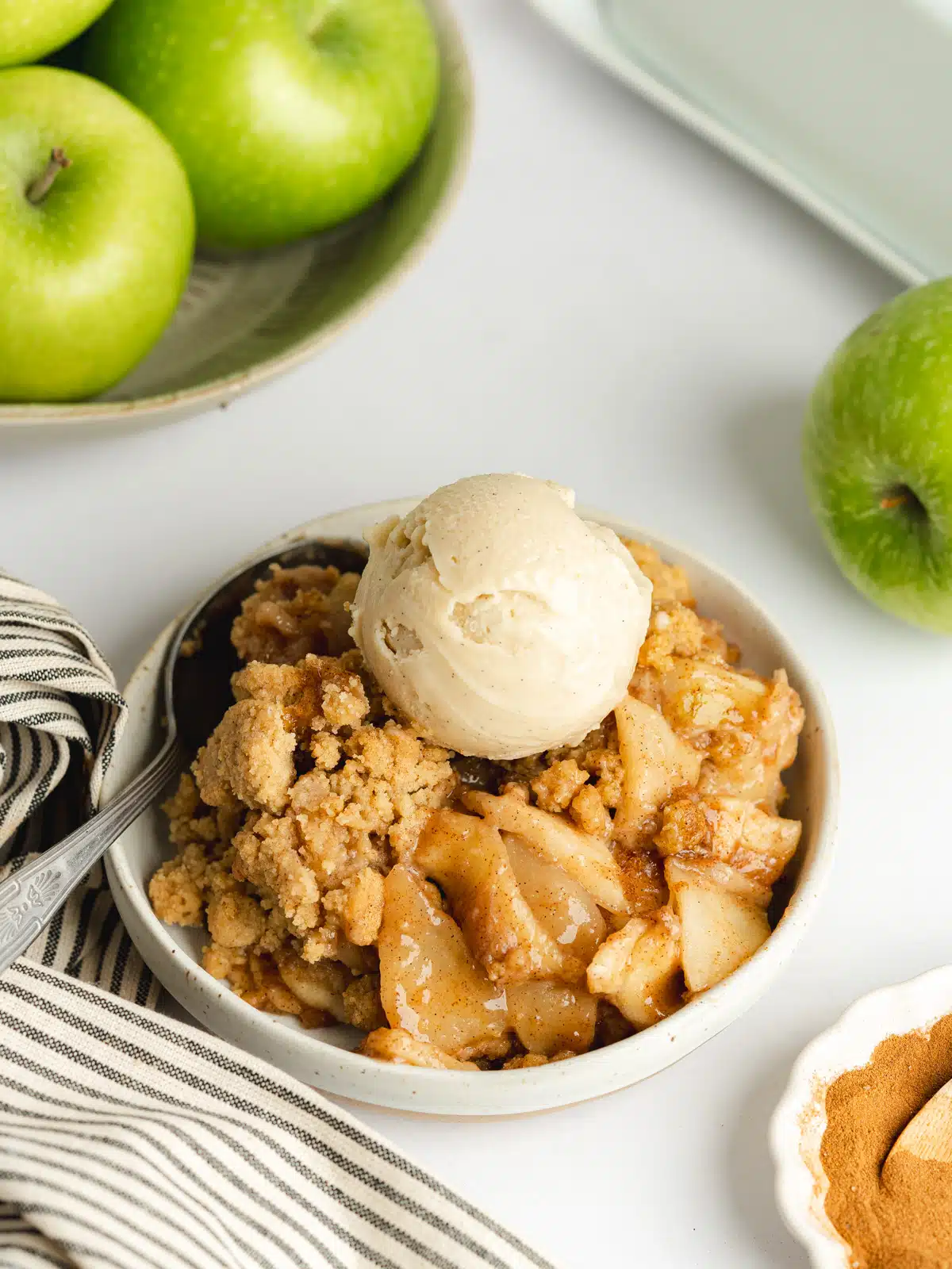 a bowl of apple crumble with a scoop of vegan vanilla ice cream and granny smith apples in the background.