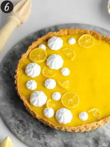 a lemon tart with lemon curd topping and piped whipped cream and small lemon slices on top of a marble cake stand.