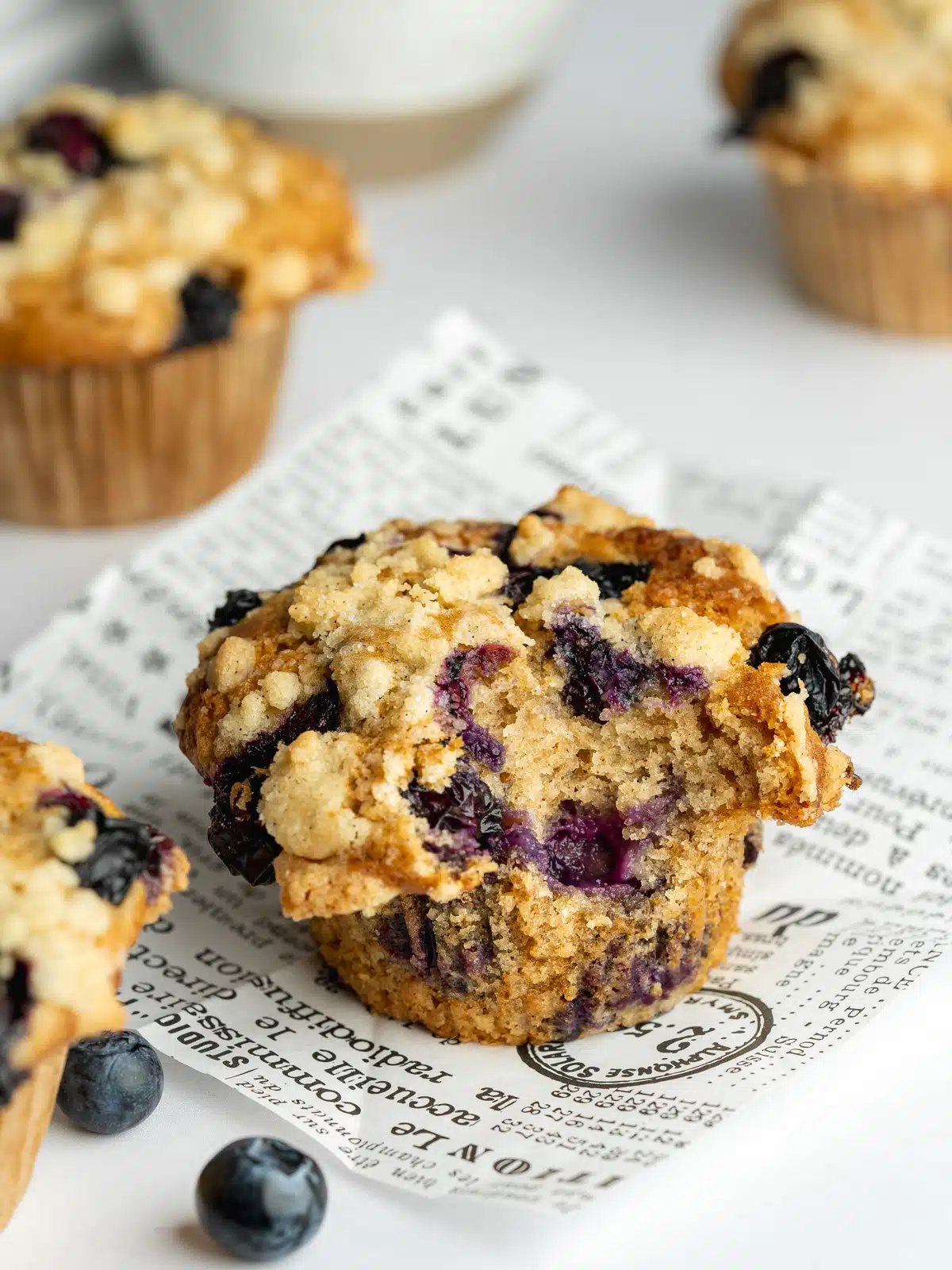 freshly baked blueberry muffins on a sheet of newspaper with a bite taken from one of them showing the moist interior.