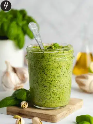 a jar of homemade basil pistachio pesto on a wooden cutting board with fresh basil, garlic, and olive oil in the background.