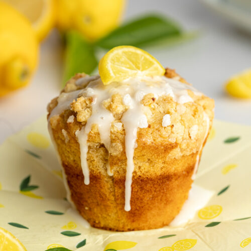 a lemon muffin with streusel topping and a small slice of lemon on top of lemon printed parchment paper.