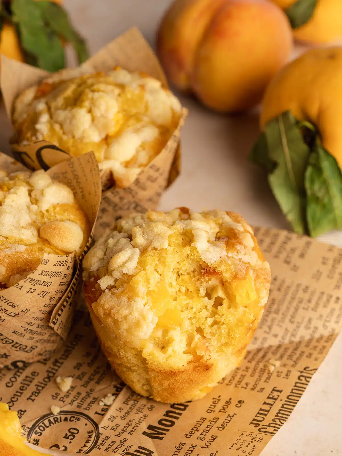 peach muffins with crumb topping on a sheet of brown newspaper with fresh peaches in the background.