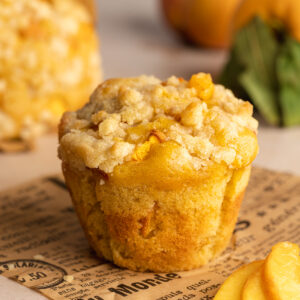 a bakery-style peach muffin with crumble topping on a piece of newspaper with fresh peaches in the background.