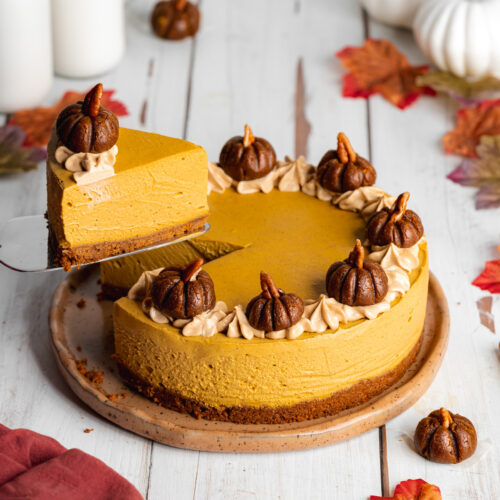 a pumpkin cheesecake topped with mini pumpkin shaped truffles with a slice being lifted away and autumn leaves scattered around it.